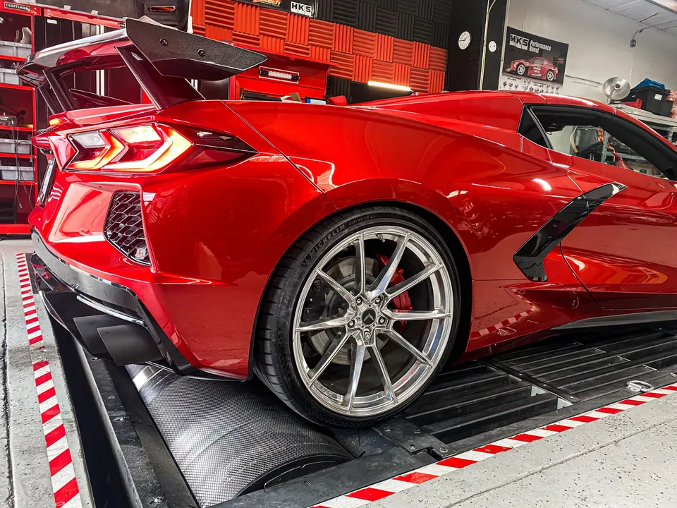 Red C8 Corvette with a large carbon fiber wing and aftermarket wheels on a dynamometer at a performance shop.