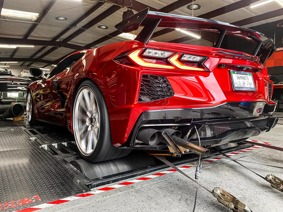 Red C8 Corvette on a dyno with custom exhaust system and large rear wing being tested at a performance shop.