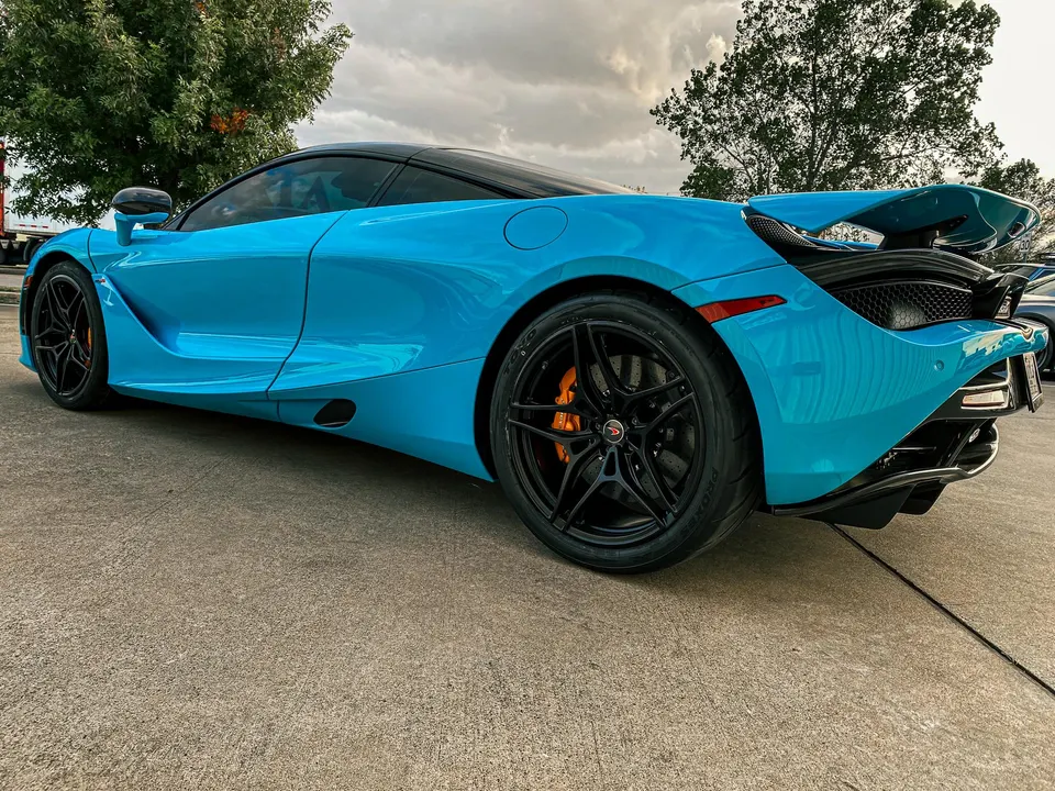 A blue McLaren 720S supercar parked outdoors, showcasing its sleek design and black wheels with orange brake calipers.