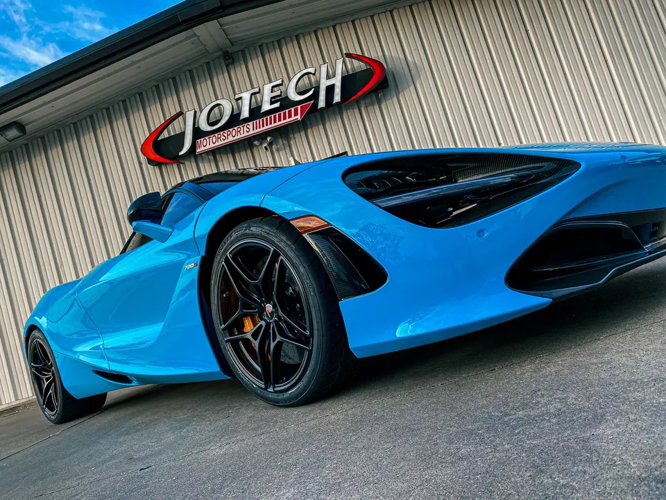 A vibrant blue McLaren 720S supercar parked at an angle in front of the Jotech Motorsports shop.