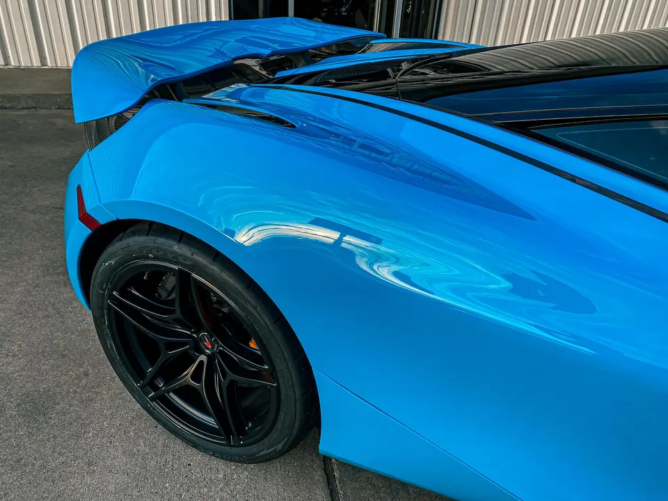 A close-up detail of a blue McLaren's rear wheel, fender, and active aero wing, showcasing its sleek design.