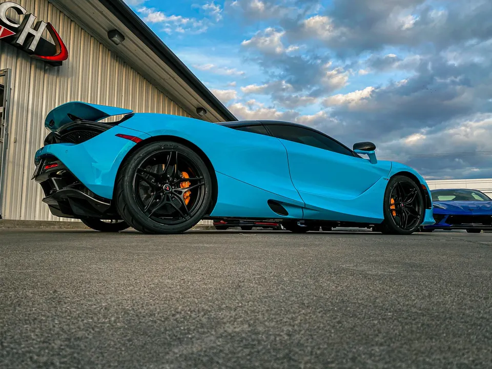 Bright blue McLaren 720S supercar parked outside Jotech Motorsports building under a cloudy sky.