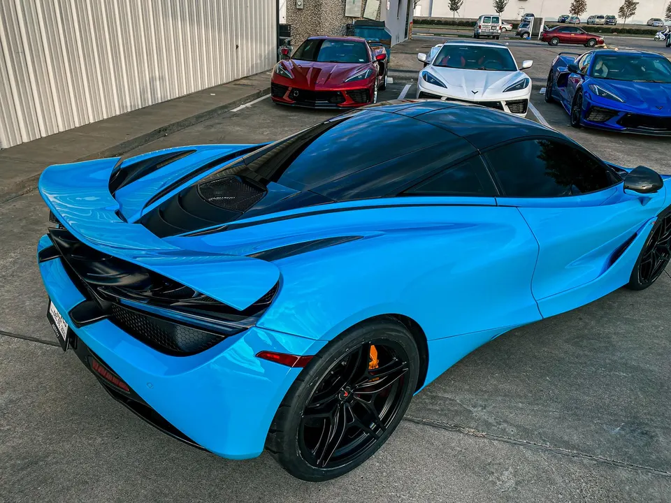 A vibrant blue McLaren 720S supercar parked in a lot, with red, white, and blue Corvette C8s visible in the background.