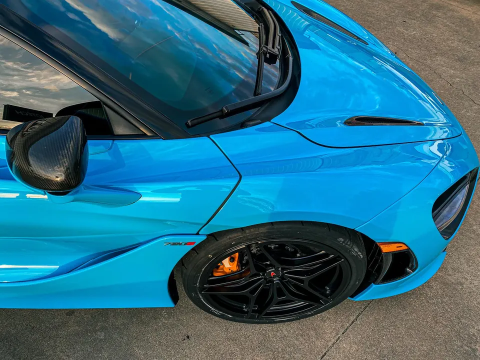 A vibrant blue McLaren 720S sports car featuring black multi-spoke wheels, bright orange brake calipers, and a carbon fiber side mirror.