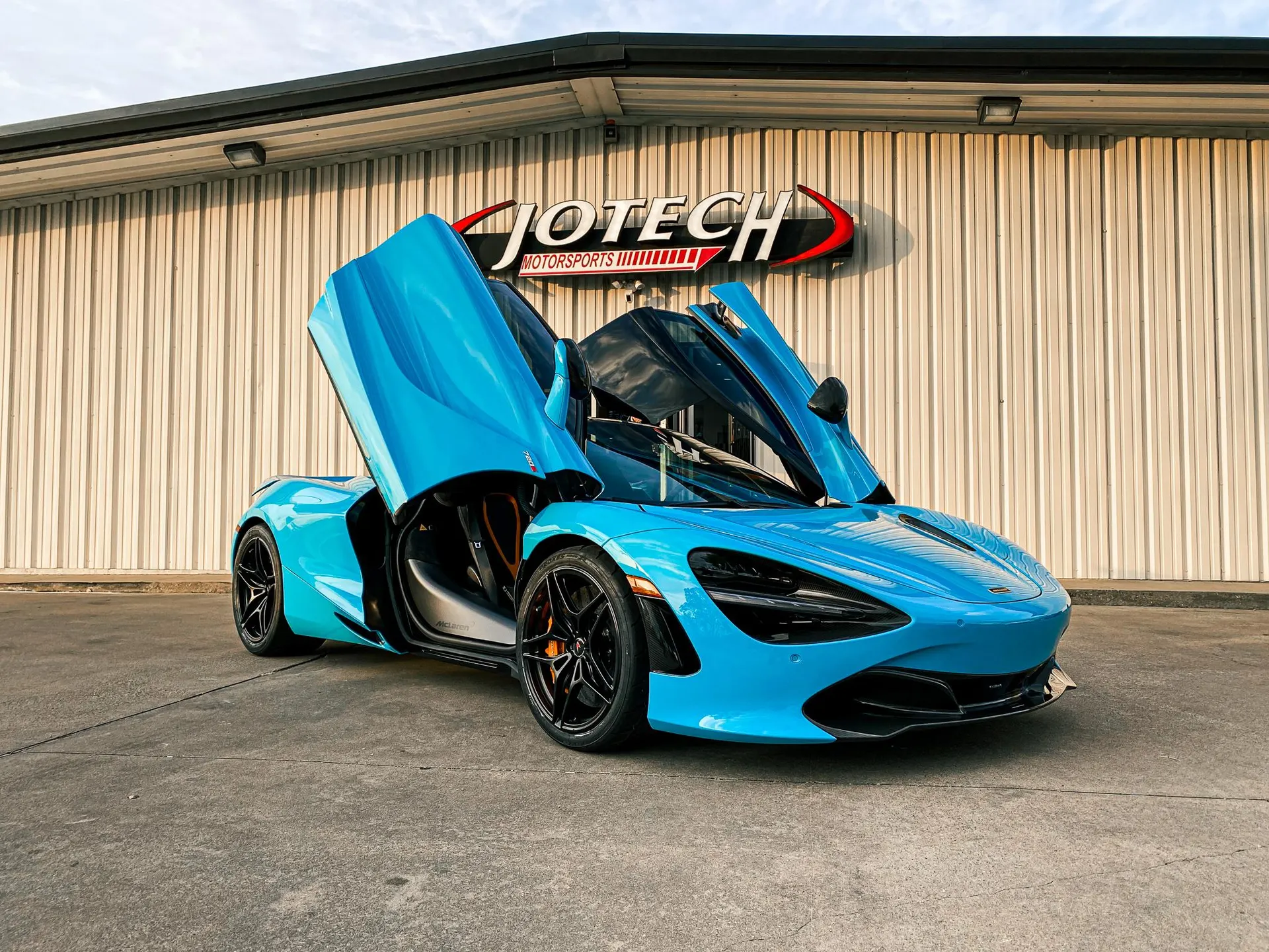 A vibrant blue McLaren supercar with its butterfly doors open, parked in front of the Jotech Motorsports shop.