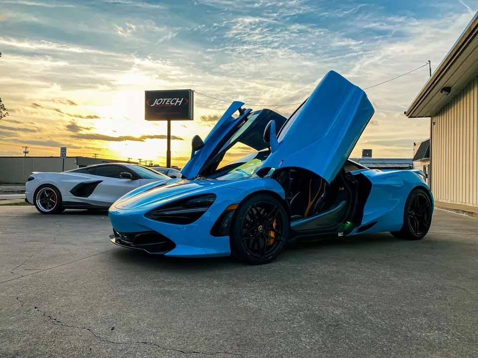 Blue McLaren 720S with butterfly doors open and a white C8 Corvette parked outside Jotech Motorsports at sunset.