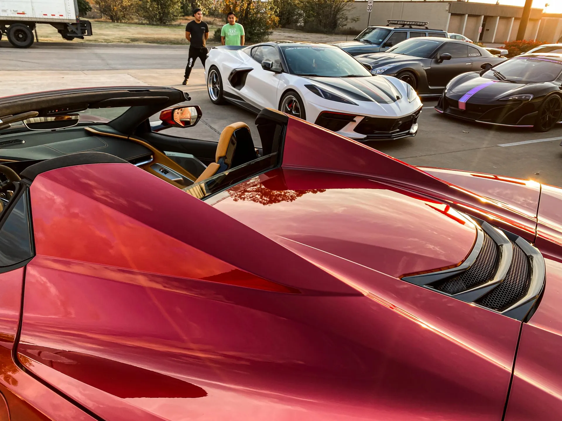 Multiple high-performance cars including a red Corvette C8, white Corvette C8, Nissan GT-R, and McLaren parked at a car meet.