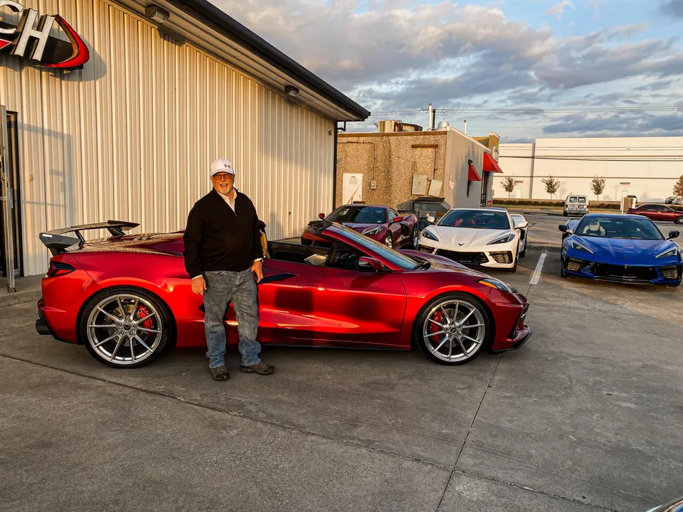 A man stands next to a red C8 Corvette convertible with a custom wing and wheels, parked outside a shop with other C8 Corvettes.