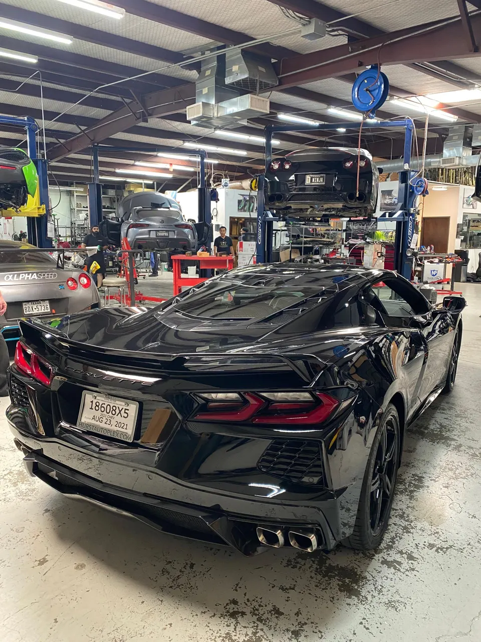 A black C8 Corvette parked in a performance tuning shop with other cars on lifts in the background.