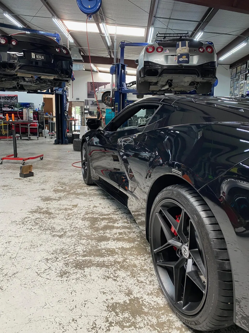 A black Corvette C8 in a performance tuning shop with two Nissan GT-Rs on lifts in the background.