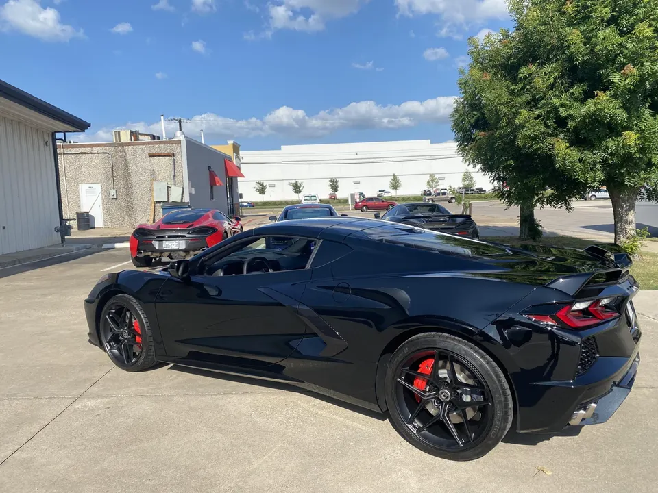 Black Chevrolet Corvette C8 parked outdoors with a red McLaren in the background on a sunny day.