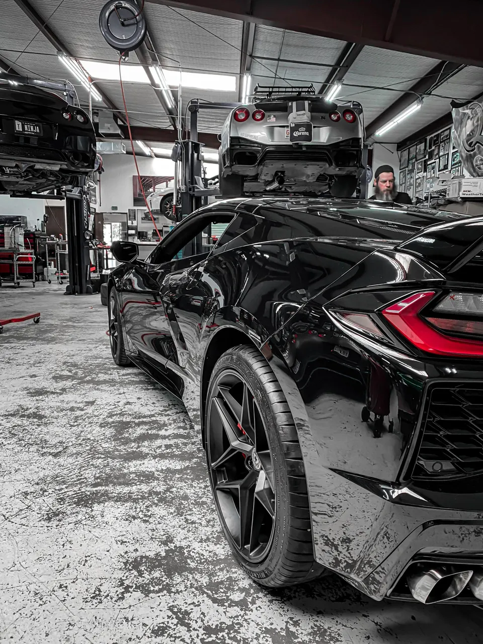 A black Corvette C8 and two Nissan GT-Rs on lifts inside a performance tuning shop with a mechanic in the background.