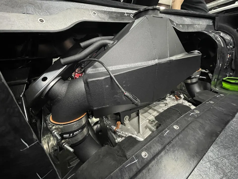 A close-up of a custom black intercooler and boost piping installed in a high-performance car's engine bay during a build.