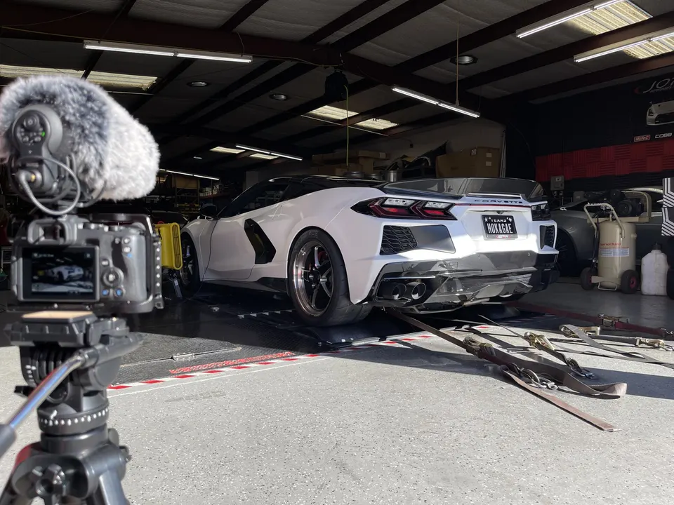 White C8 Corvette strapped to a dyno for performance tuning, with a camera recording the session in a shop.