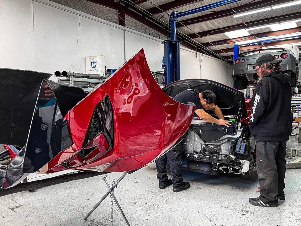 Mechanics working on a red Corvette C8 with its rear bumper removed, exposing the exhaust, in a performance shop.