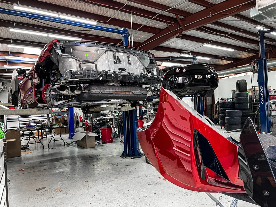 A red C8 Corvette with its rear bumper removed on a lift in a performance shop, exposing the exhaust system. Another black car is on a lift in the background.