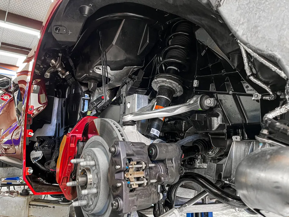 A red Corvette C8 on a lift in a shop, with the rear wheel removed, showing the suspension, red brake caliper, and axle.