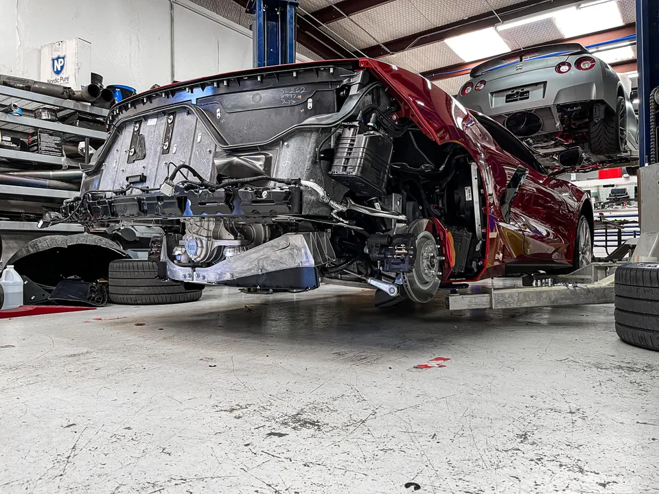 Red Corvette C8 undergoing disassembly on a lift in a performance shop, with a Nissan GT-R on a lift in the background.