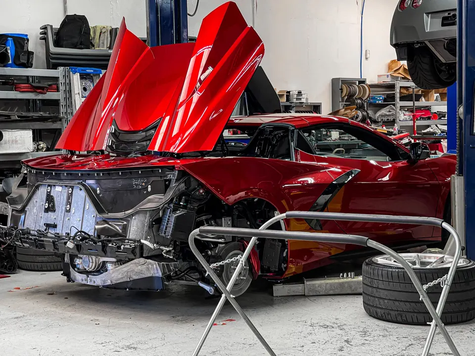 Red Chevrolet Corvette C8 undergoing disassembly in a performance tuning shop, with its front bumper removed and hood open.