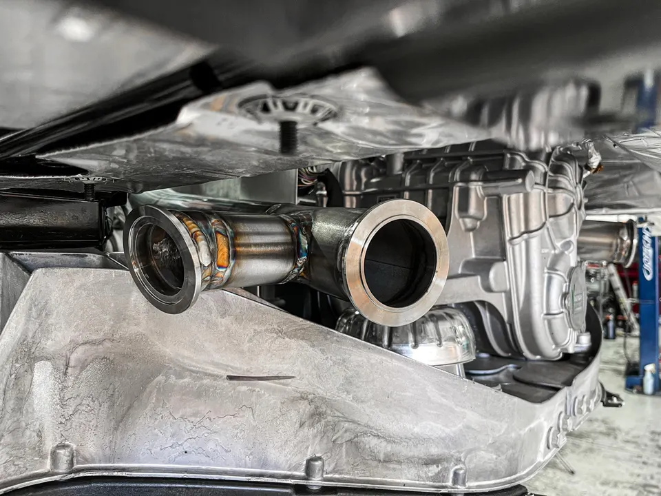 Close-up of a custom stainless steel exhaust downpipe section with visible welds being installed on a performance car.
