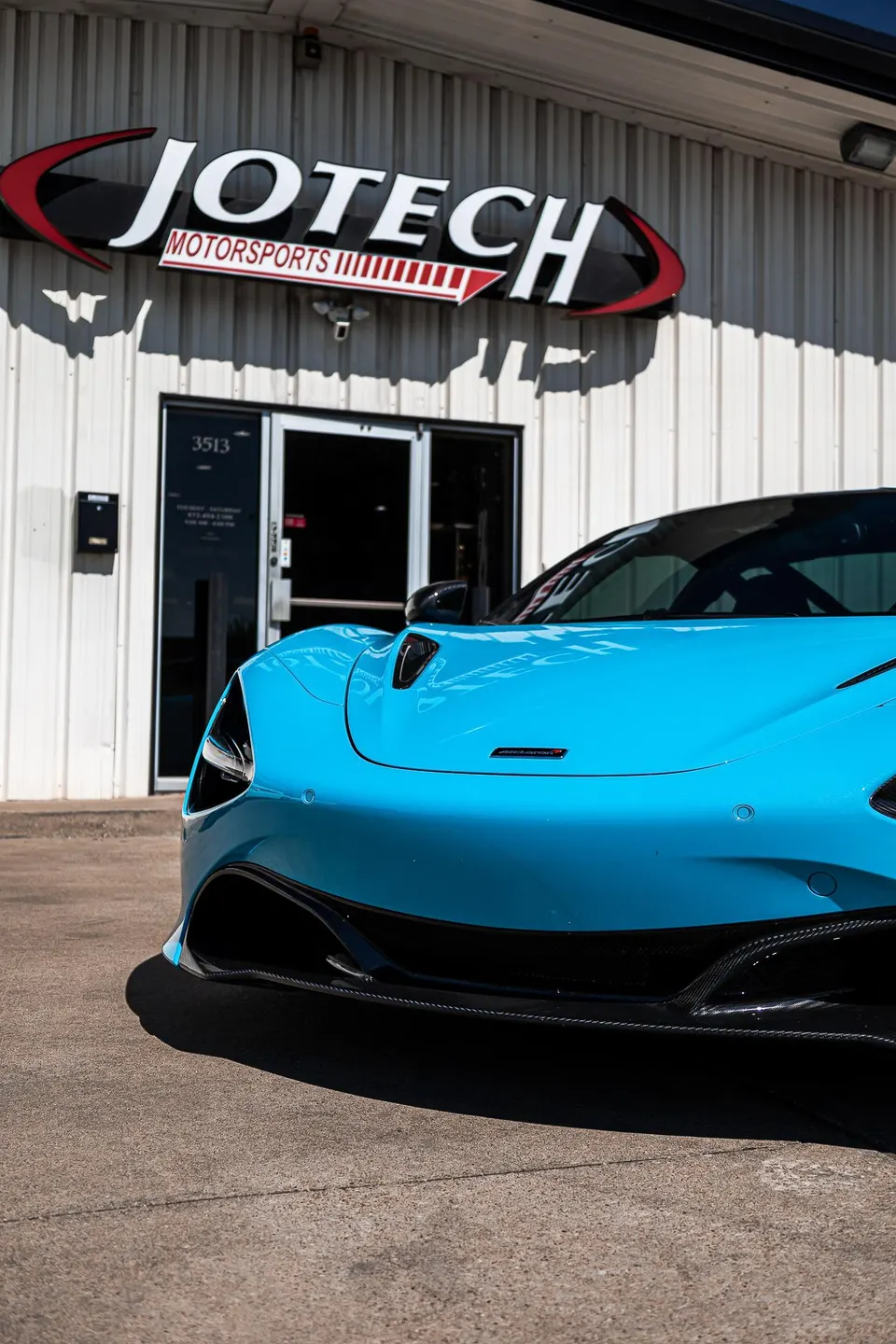 A vibrant blue McLaren supercar parked outside the Jotech Motorsports facility, showcasing the shop's storefront and signage.