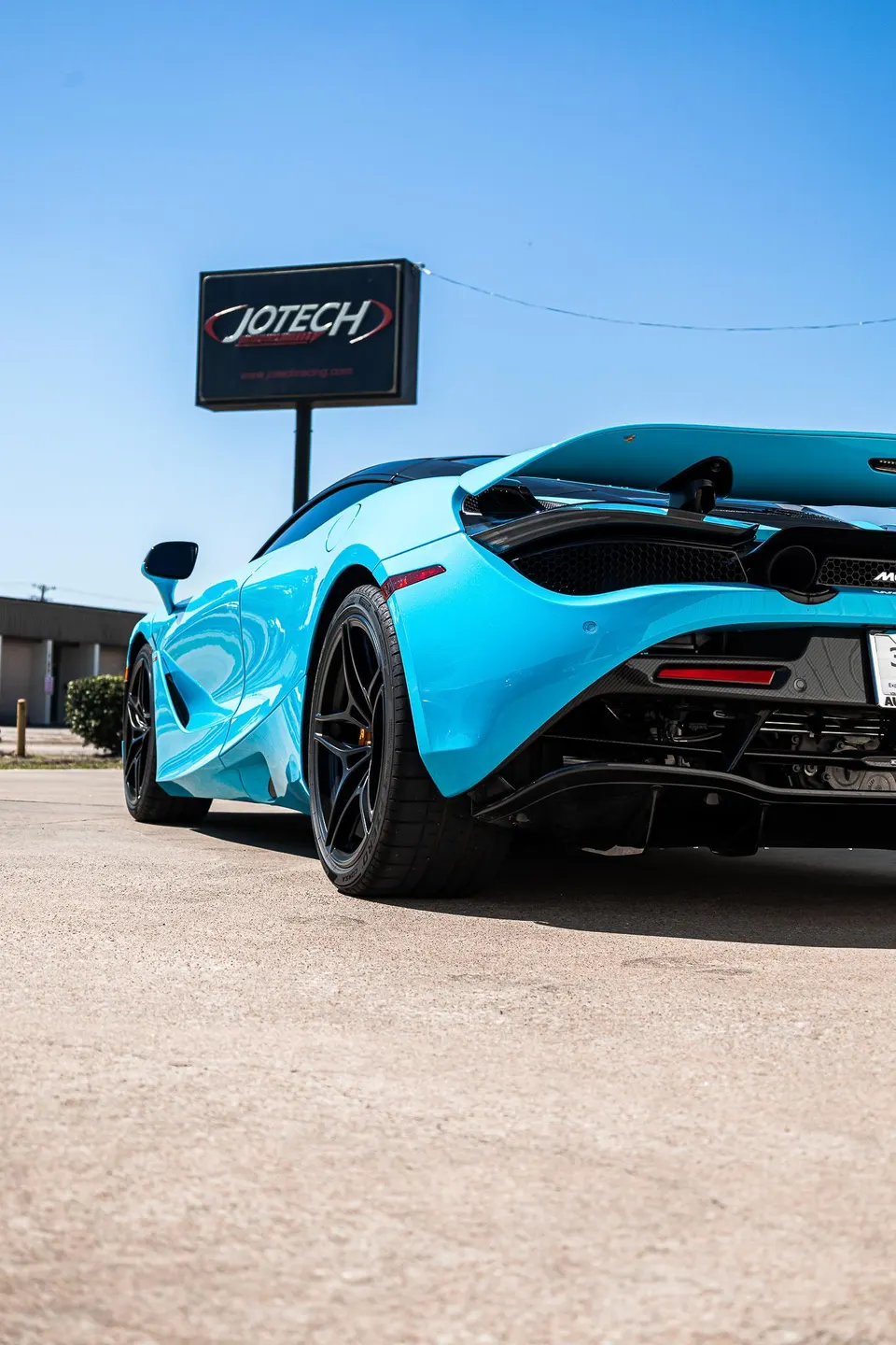 A vibrant blue McLaren 720S supercar parked outdoors, showcasing its aggressive rear design with a carbon fiber diffuser and active rear wing.