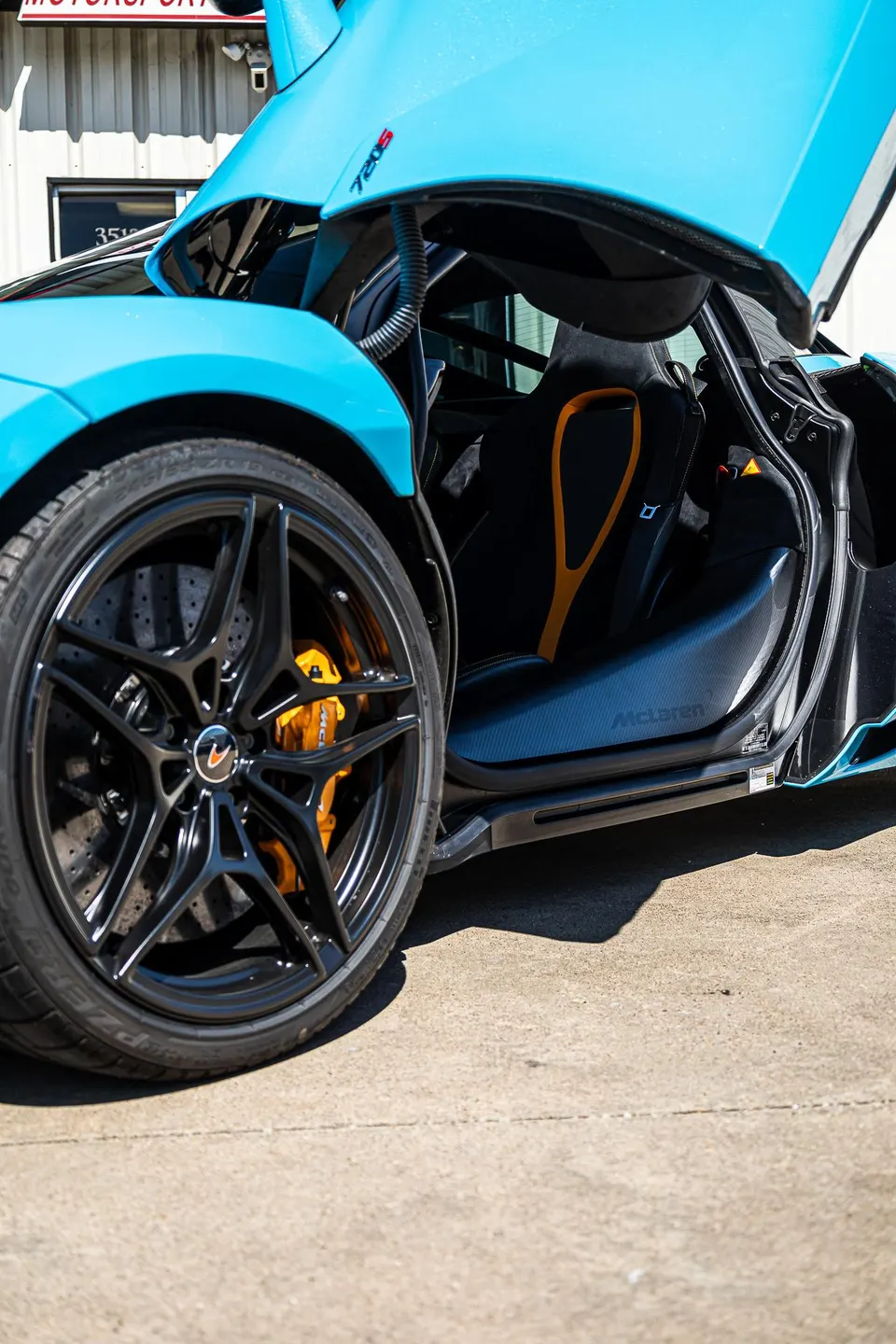 A blue McLaren 720S with its door open, showcasing black multi-spoke wheels, orange brake calipers, and carbon fiber interior trim.