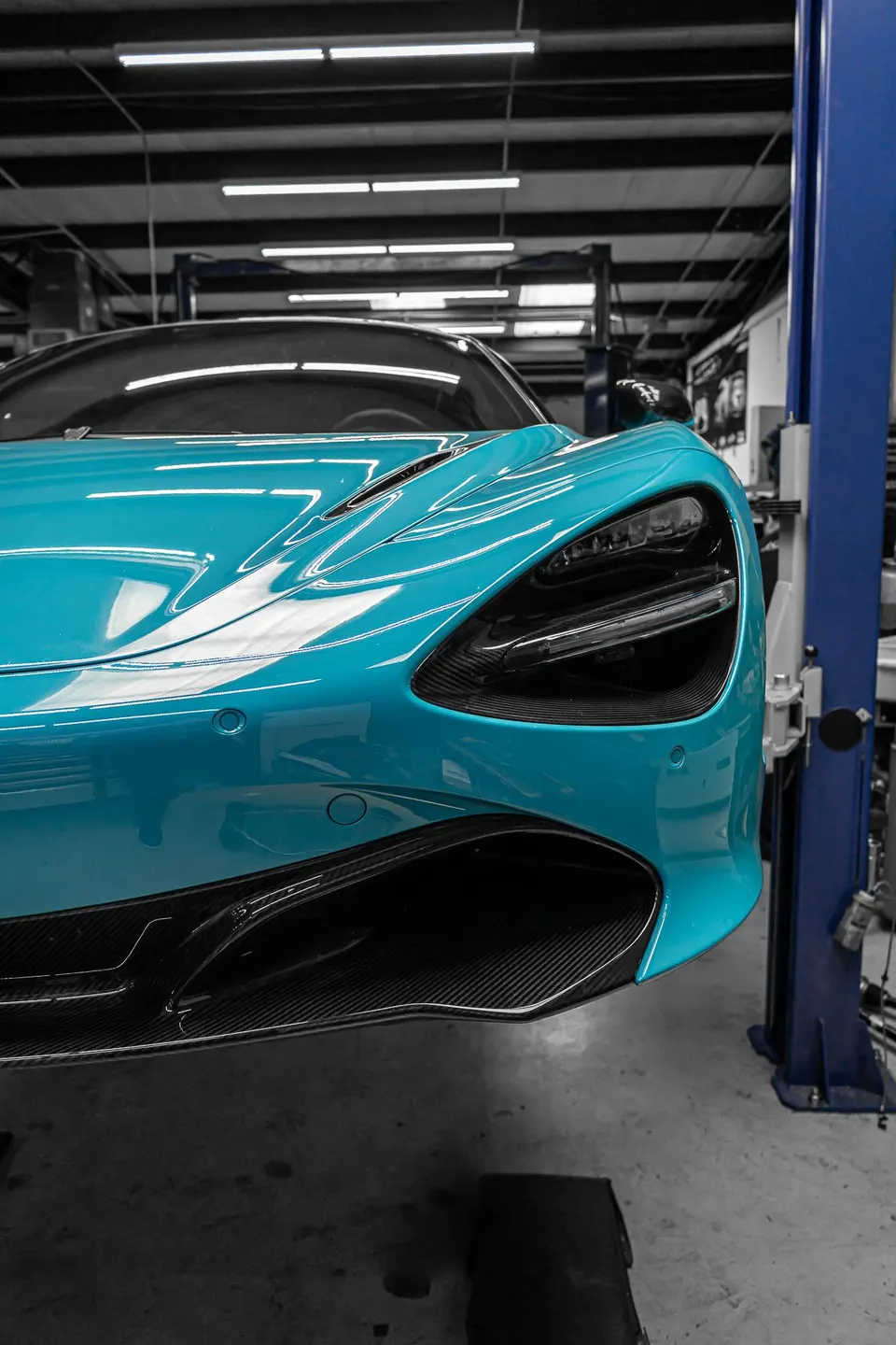 A blue McLaren 720S on a lift in a performance tuning shop, showcasing its front bumper and carbon fiber aero.