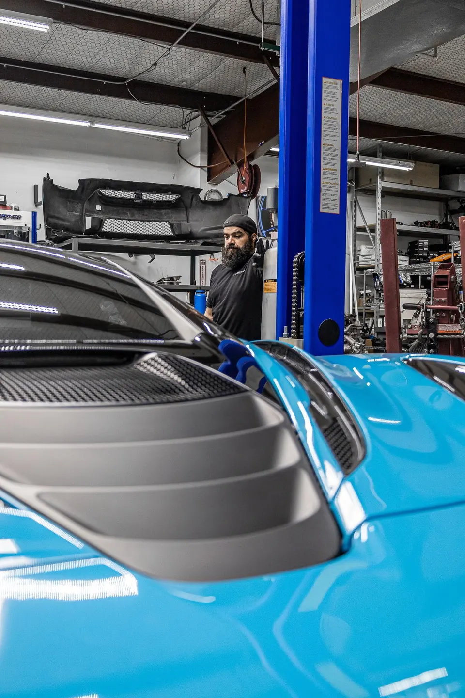 Blue McLaren supercar in a performance tuning shop with a mechanic and car lift in the background.