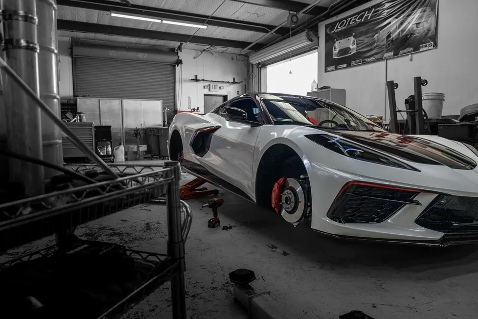 A white Corvette C8 on jacks in a performance shop, with red brake calipers visible and wheels removed for service.