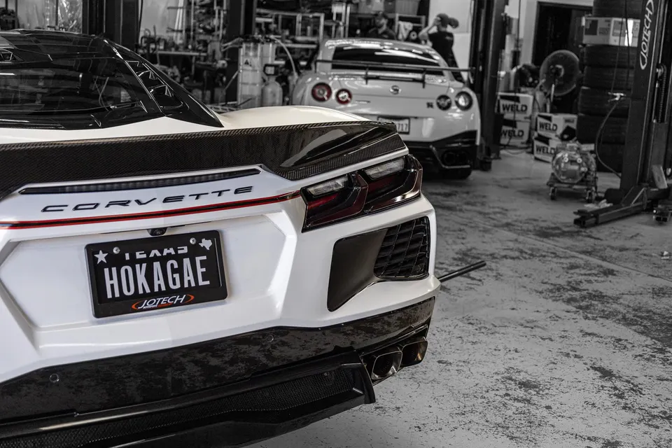 White Corvette C8 rear view with carbon fiber spoiler and diffuser, parked in a performance tuning shop with a Nissan GT-R in the background.
