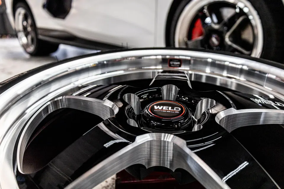 Close-up of a black and silver Weld wheel with a white car in the blurred background, showcasing the wheel's design.