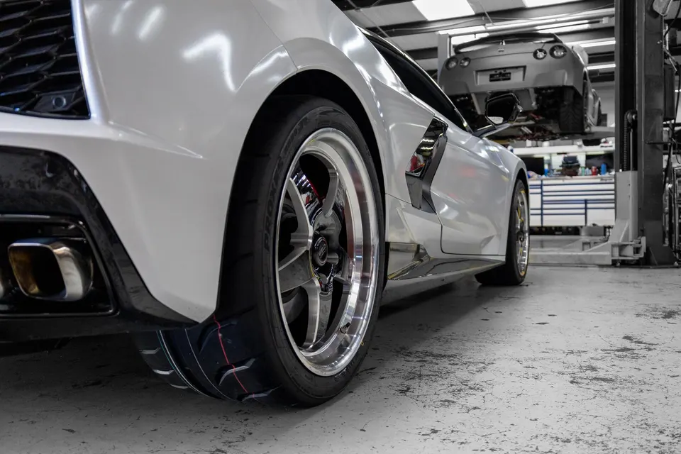 A white C8 Corvette with aftermarket drag wheels and tires in a performance tuning shop, with a Nissan GT-R on a lift.