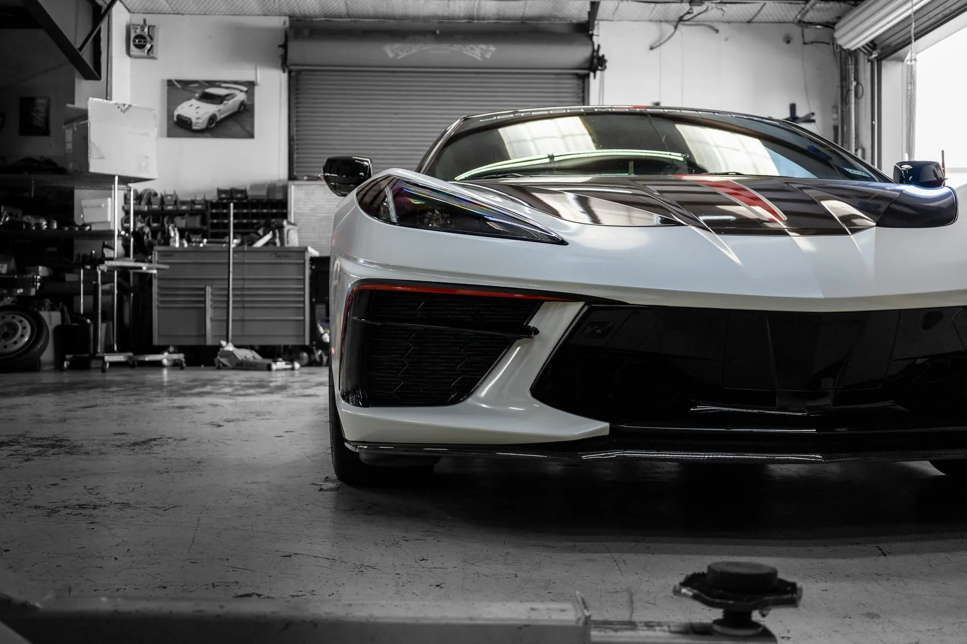 A white C8 Corvette with black and red racing stripes on the hood, parked in a garage with tools in the background.