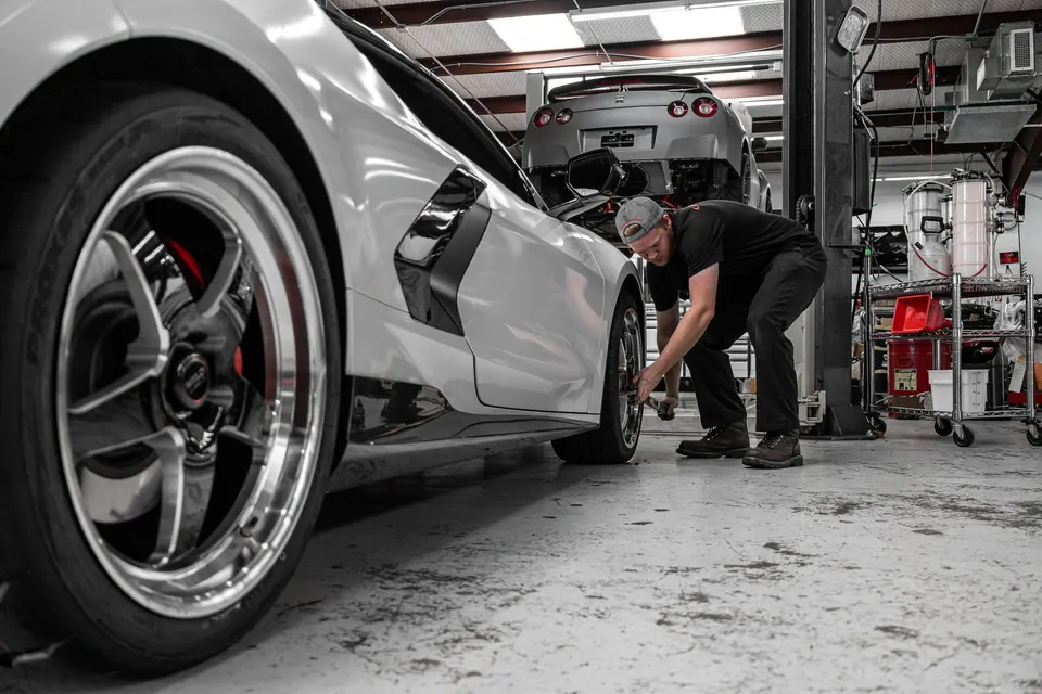 A mechanic works on the wheel of a white Corvette C8 in a performance tuning shop, with a Nissan GT-R on a lift in the background.
