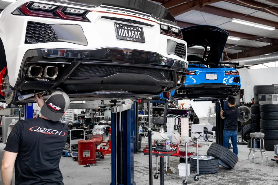 Two C8 Corvettes on lifts in a performance shop, with mechanics working on the exhaust system of a white Corvette.