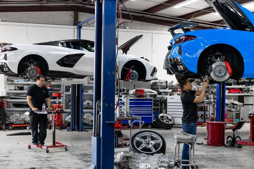 Mechanics working on multiple Chevrolet Corvette C8s on lifts in a performance tuning shop, with wheels and a transmission on the floor.
