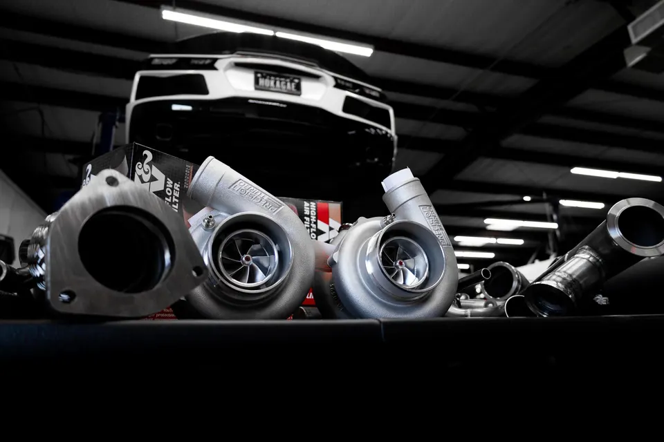 Precision turbochargers and exhaust components laid out in a performance shop with a white sports car on a lift in the background.