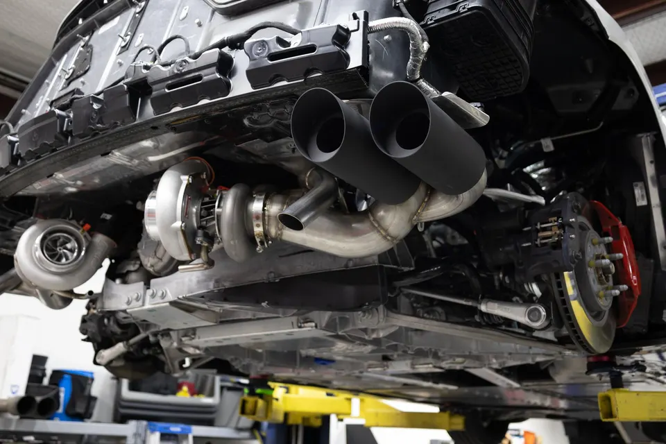 Underside view of a performance car on a lift, showcasing a twin-turbo setup, custom exhaust system, and red brake calipers.