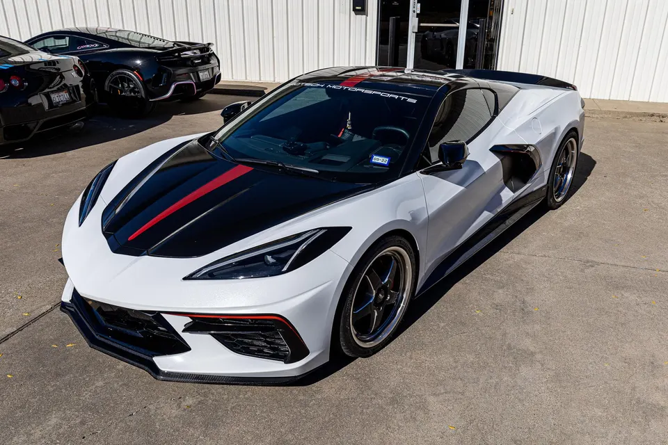 White Chevrolet Corvette C8 with black and red racing stripes, carbon fiber aero, and aftermarket wheels parked outside Jotech Motorsports.