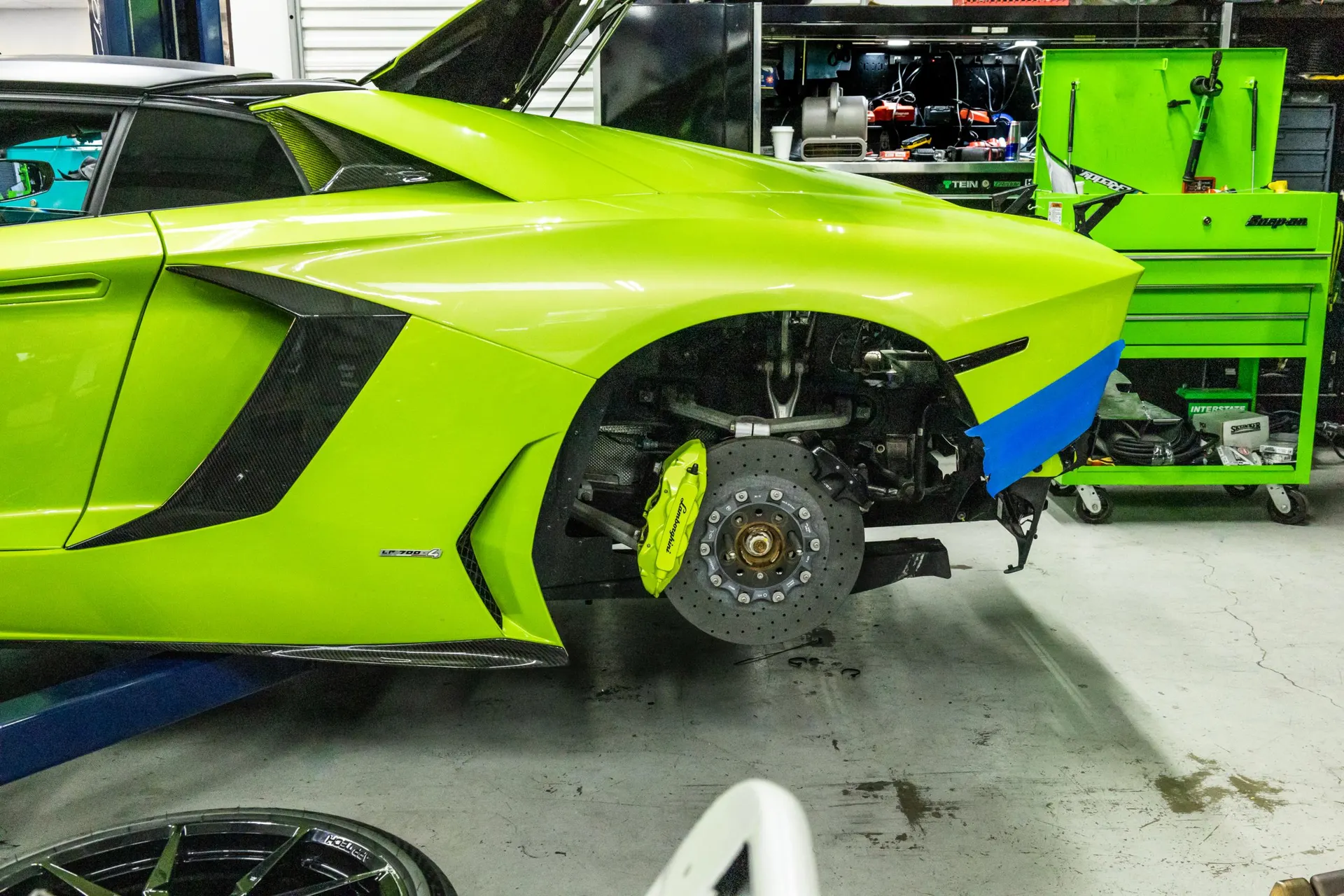 A lime green Lamborghini Aventador in a shop with the rear wheel removed, showcasing the brake system and suspension.