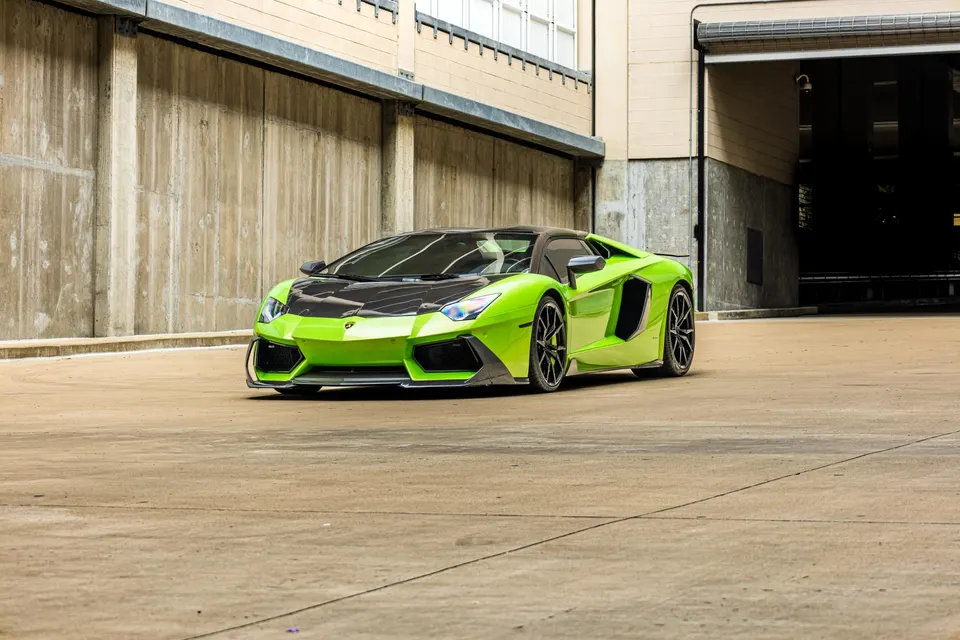 A vibrant green Lamborghini Aventador with a black carbon fiber hood parked in an urban industrial setting.