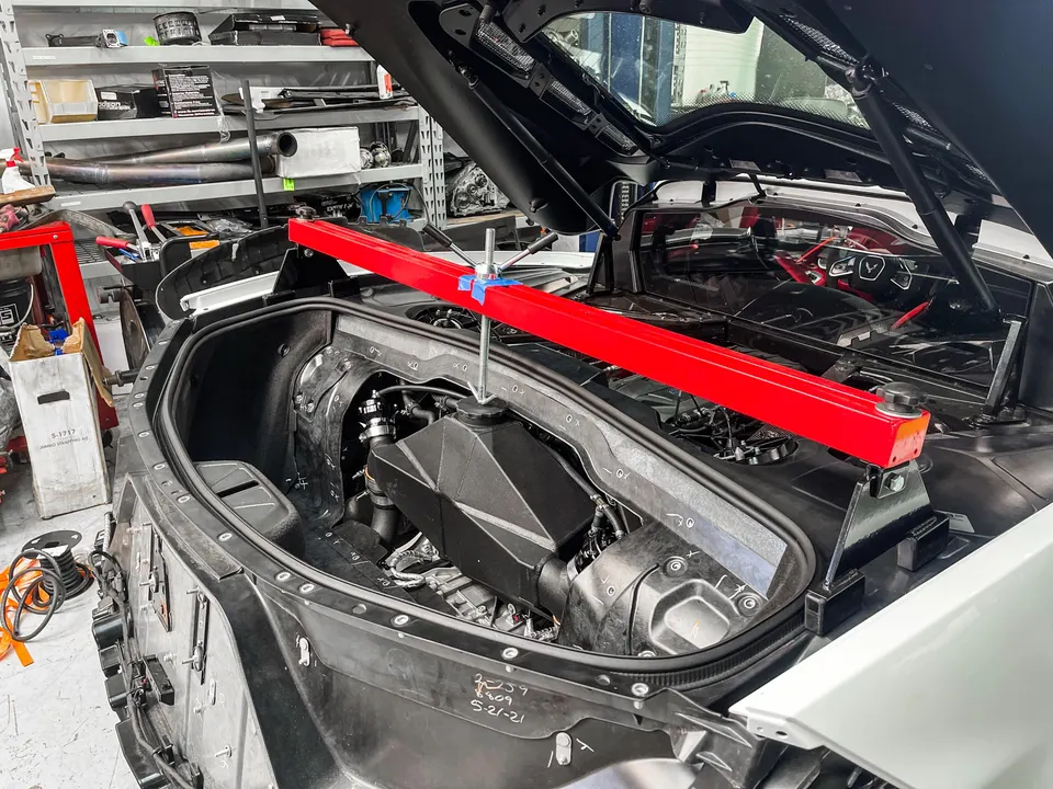 A white Corvette C8 with its rear open, revealing the mid-engine bay, supported by a red bar in a Jotech Motorsports shop.
