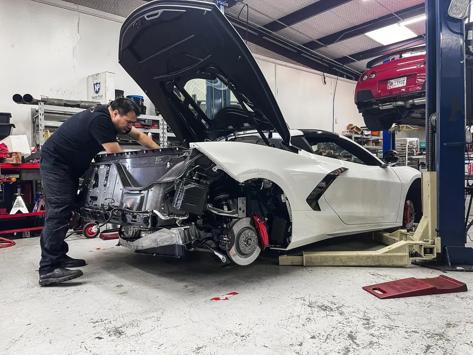Mechanic working on a white C8 Corvette with its rear bumper removed in a performance tuning shop.
