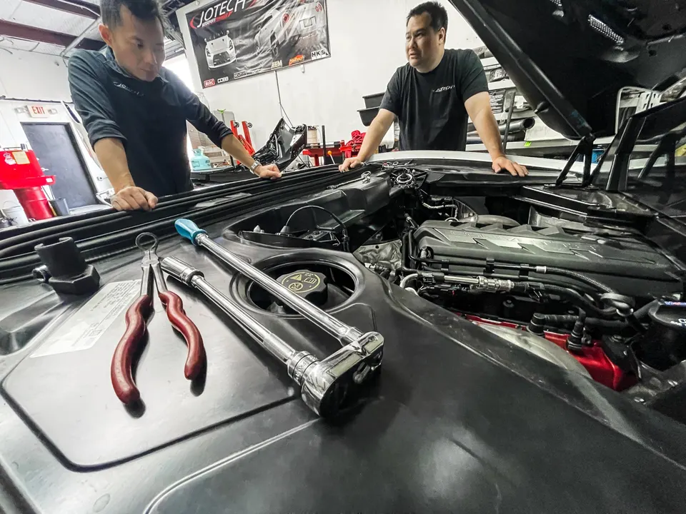 Two mechanics inspecting the engine bay of a Corvette C8 at Jotech Motorsports shop with tools on the engine cover.