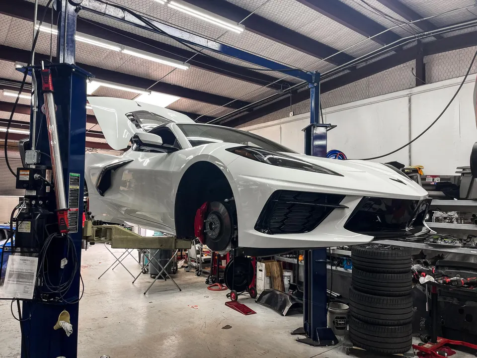 A white C8 Corvette on a lift in a performance tuning shop with its wheels removed and engine cover open.