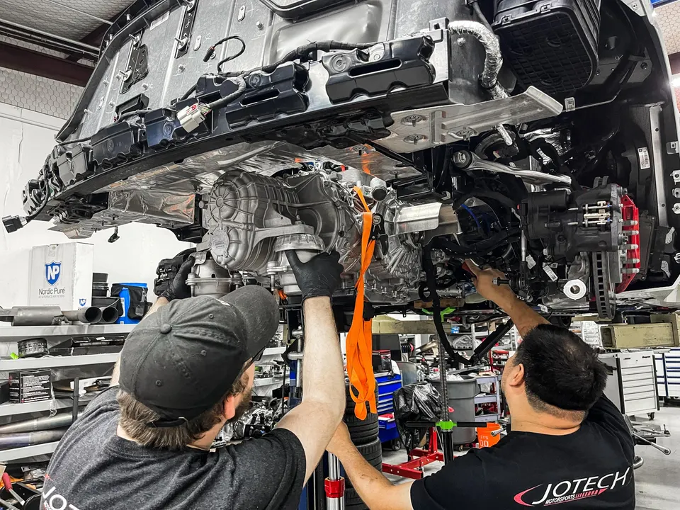 Mechanics at Jotech Motorsports installing a transmission on a car lifted in the shop.