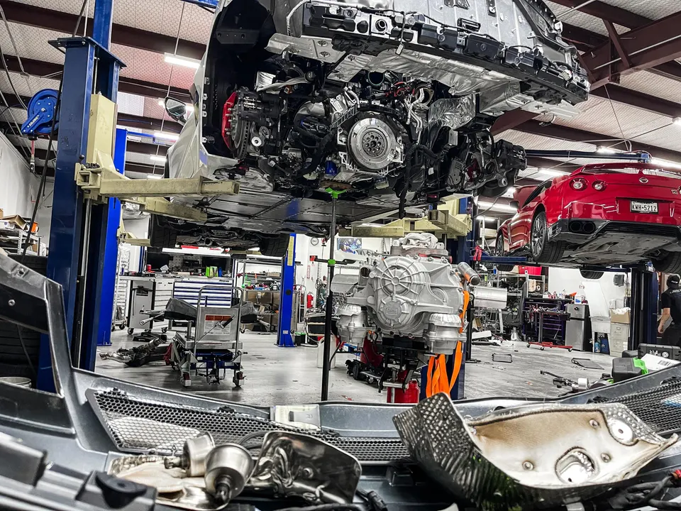 A busy performance tuning shop with a C8 Corvette on a lift undergoing engine removal, a red Nissan GT-R on another lift, and a detached powertrain on the floor