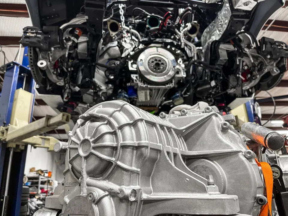 A car on a lift in a shop with its engine bay exposed, and a silver transmission housing in the foreground.