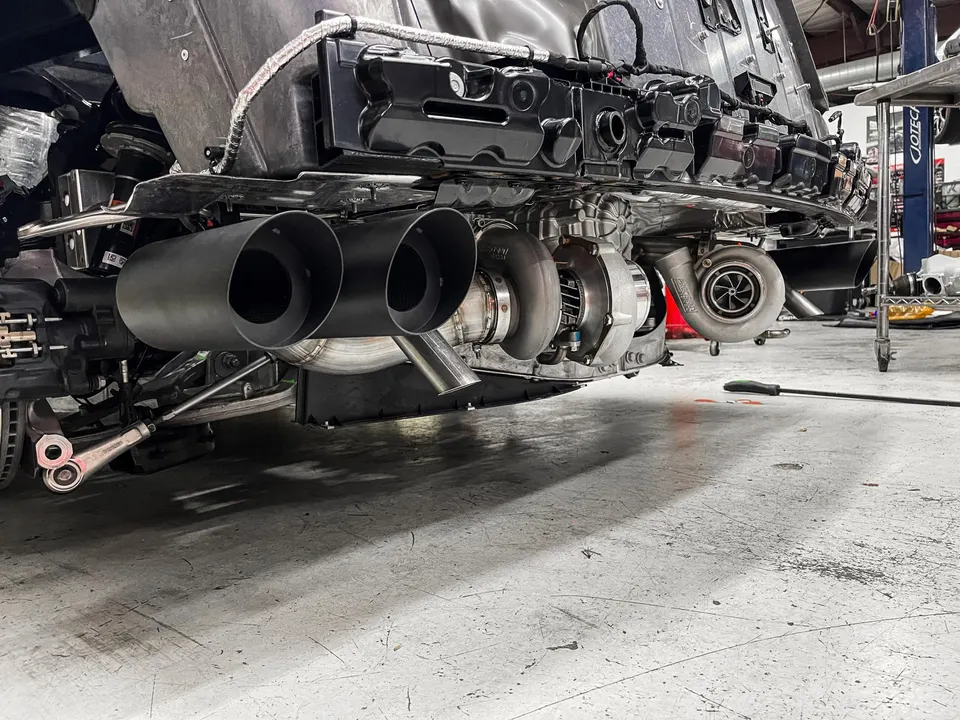 Underside view of a performance car in a shop, featuring twin turbochargers and a custom exhaust system installation.